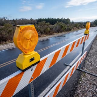 Illustration: a solar amber LED barricade light, switched off, on a barricade closing an access road in daylight.