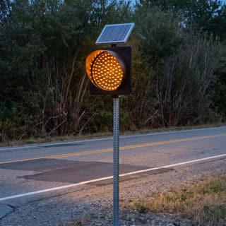 Illustration: a solar amber LED flashing beacon in a black plastic housing glowing beside a dip in a country road after dark.