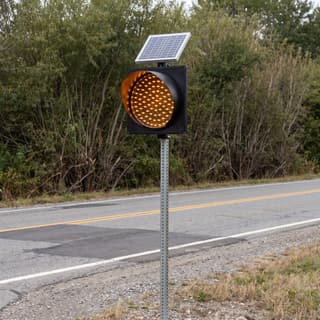 Illustration: a solar amber LED flashing beacon in a black plastic housing on a post beside a dip in a country road.