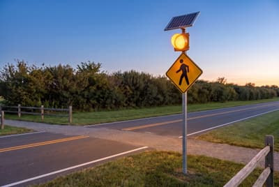 Illustration: a solar amber flashing beacon above a pedestrian warning sign where a trail crosses a rural road after dark.