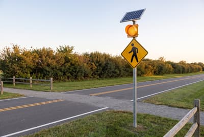 Illustration: a solar amber flashing beacon mounted above a pedestrian warning sign where a trail crosses a rural road.