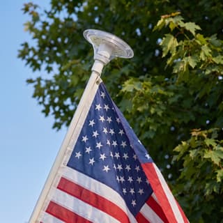 Illustration: a solar top-mount LED light, switched off in daylight, on the tip of a residential flag pole above an American flag.