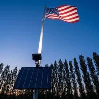 Illustration: a solar LED flag pole light with a large solar panel uplighting an American flag at dusk.