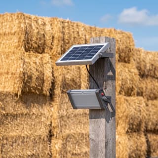 Illustration: a small solar LED flood light, switched off in daylight, with its separate panel on a timber post in a farmyard.