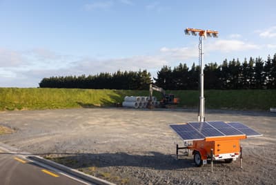 Illustration: a trailer-mounted solar light tower with its panels charging in a construction yard in daylight.