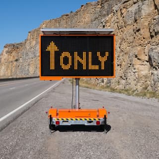 Illustration: a solar full-matrix LED message sign trailer showing an up arrow and ONLY on a gravel shoulder in a highway rock cut.