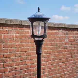 Illustration: a black solar LED lantern on a decorative pole, switched off in daylight, in front of a brick garden wall.