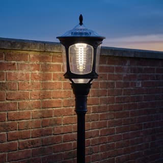 Illustration: a black solar LED lantern on a decorative pole lit in front of a brick garden wall at dusk.