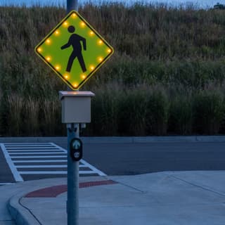 Illustration: a solar LED-bordered pedestrian crossing sign with a push button glowing at a marked crossing on a municipal parking lot drive after dark.