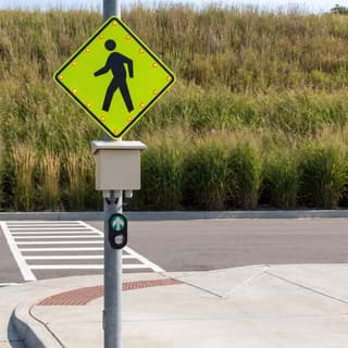 Illustration: a solar LED-bordered pedestrian crossing sign with a push button at a marked crossing on a municipal parking lot drive.