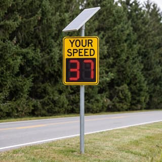Illustration: a solar LED radar speed sign showing a driver's speed in red on a two-lane county road.