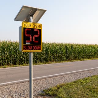 Illustration: a solar LED radar speed sign showing a speed and a lit SLOW DOWN message on a county highway.