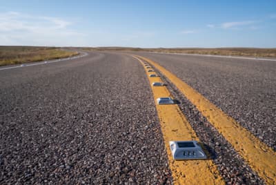 Illustration: solar LED road studs set along the centre and edge lines of a rural road in daylight.