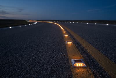 Illustration: solar LED road studs tracing the centre and edge lines of an unlit rural road at night.