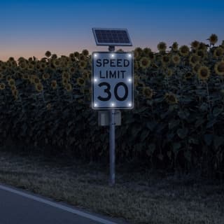 Illustration: a solar LED flashing 30 mph speed limit sign glowing on a country road beside a sunflower field after dark.