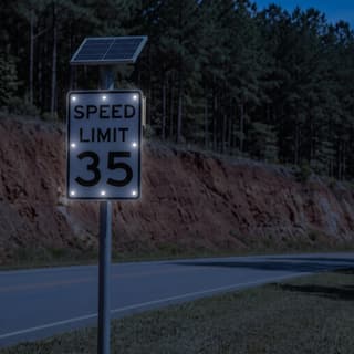 Illustration: a solar LED flashing 35 mph speed limit sign glowing on a state road below a red clay bank after dark.