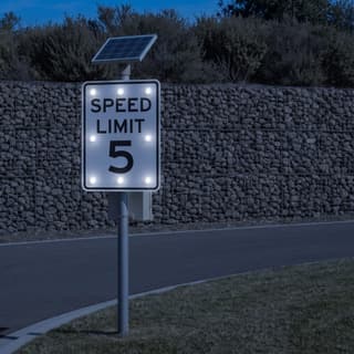 Illustration: a solar LED flashing 5 mph speed limit sign glowing on a park drive in front of a stone gabion wall after dark.