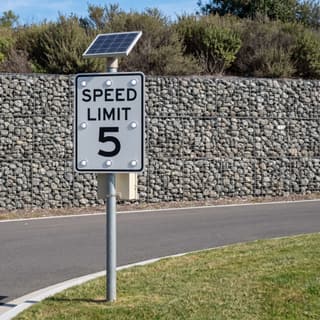 Illustration: a solar LED flashing 5 mph speed limit sign on a park drive in front of a stone gabion wall.