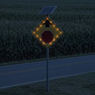 Illustration: a solar LED flashing stop ahead sign glowing on a country road beside standing corn after dark.