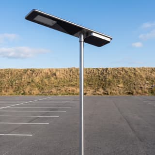 Illustration: a glossy black all-in-one solar LED lot light, switched off and charging, over a rural park-and-ride lot in daylight.