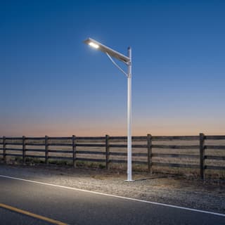 Illustration: a small solar LED light side-mounted on a pole lighting a gravel farm-lane turn-in at dusk.