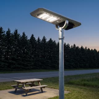 Illustration: a compact silver all-in-one solar LED light on a low pole over a highway picnic pull-off at dusk.