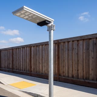 Illustration: a silver all-in-one solar LED street light, switched off and charging, over a residential sidewalk and kerb ramp in daylight.