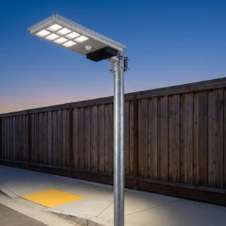 Illustration: a silver all-in-one solar LED street light on a pole lighting a residential sidewalk and kerb ramp at dusk.