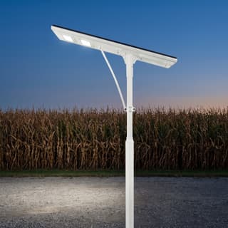 Illustration: a white all-in-one solar LED street light capping its pole over a fairgrounds gravel overflow lot at dusk.
